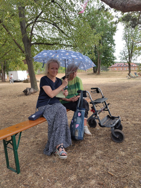 Abendgottesdienst bei der Umweltstation: Zwei Gottesdienstbesucherinnen sitzen unter einem Regenschirm und freuen sich über den kurzen Regenschauer.